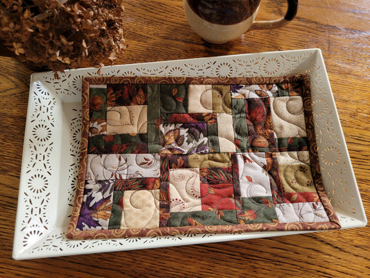 A square quilted patchwork placemat with autumn and floral patterns, displayed on a white decorative tray on a wooden table.