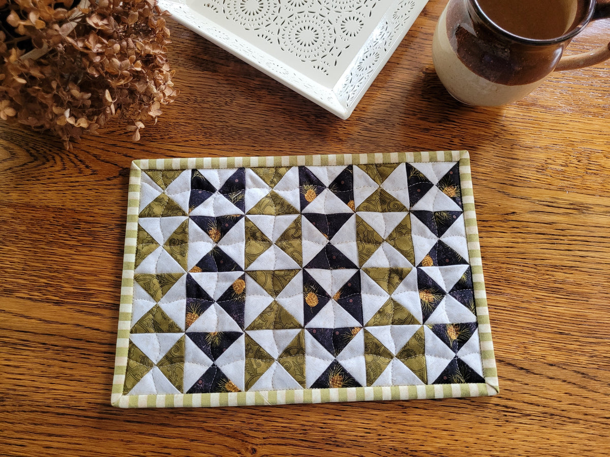 A small rectangular quilted fabric placemat with a geometric triangle pattern in olive green, white, and black, featuring a striped border, displayed on a wooden table next to a ceramic mug and a white decorative tray.