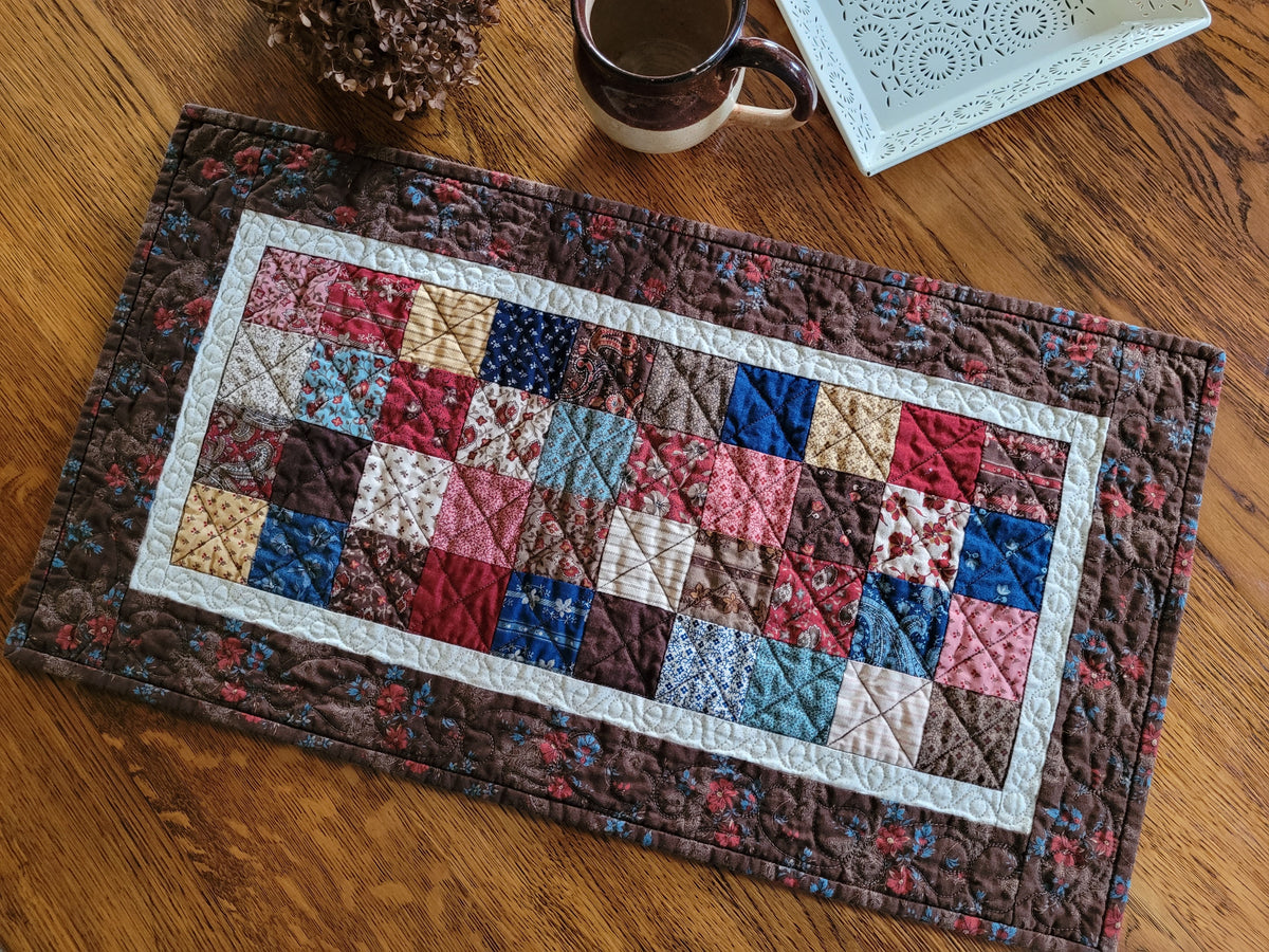 A rectangular quilted table runner with a patchwork design in earthy tones and floral fabrics, styled on a wooden tabletop with a mug, dried flowers, and decorative plate nearby.
