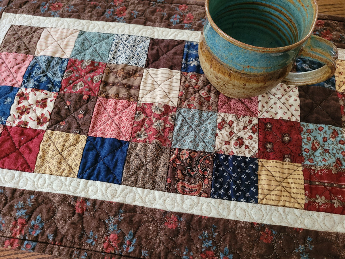 A close-up of a handmade quilted table runner with a rustic pottery mug placed on top. The table runner features a patchwork design of various fabric squares in red, blue, brown, beige, and floral patterns, bordered by thick white quilted stripe.