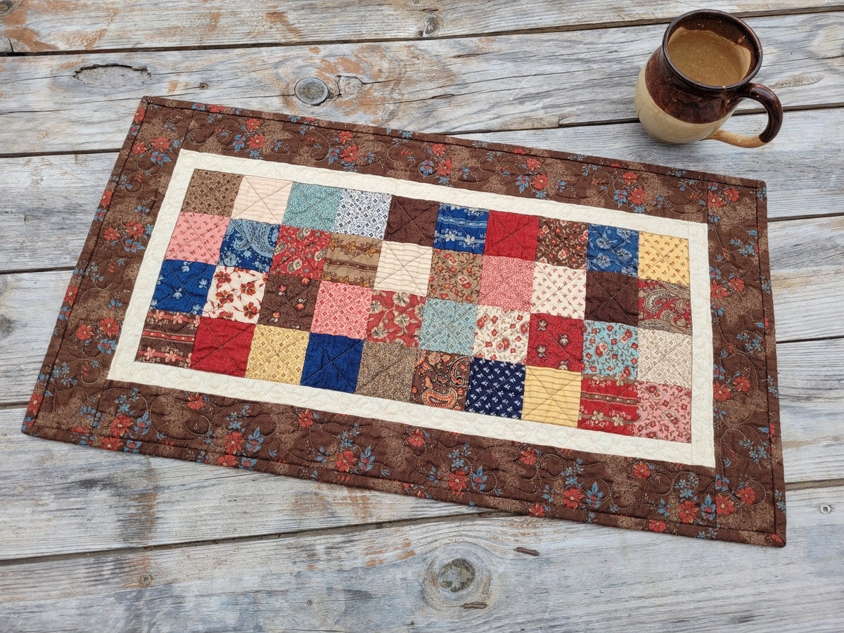 A handmade quilted table runner with a colorful patchwork center and a brown floral border, displayed on a rustic gray wooden table next to a ceramic mug.