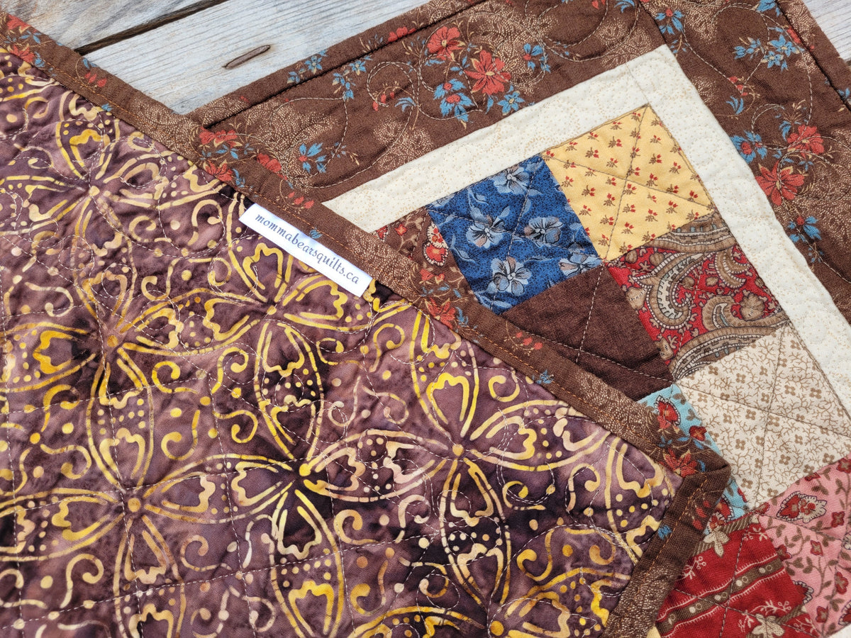 A close-up of a quilted table runner with patterned fabrics in brown, gold, blue, red, and cream, laid over a light wood surface. The runner is folded to show the back and with small fabric tag visible near the center.