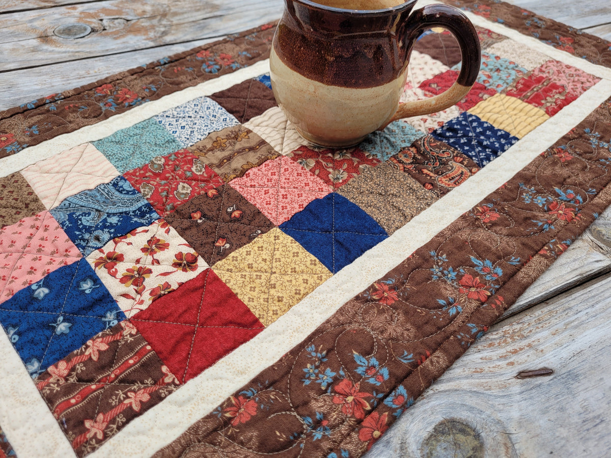 A handmade quilted patchwork table runner with a rustic floral and geometric pattern, styled on a weathered wooden table with a ceramic mug resting on top.