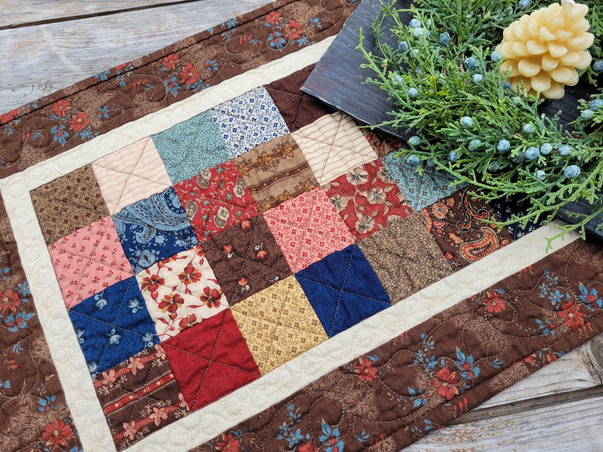 A quilted patchwork table runner with a rustic floral design, styled on a wooden tabletop with greenery and a pinecone beeswax candle.
