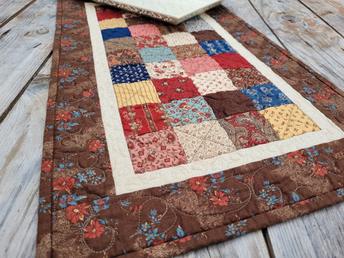 A handmade quilted table runner with a patchwork center and floral brown borders, laid out on a rustic gray wooden table.