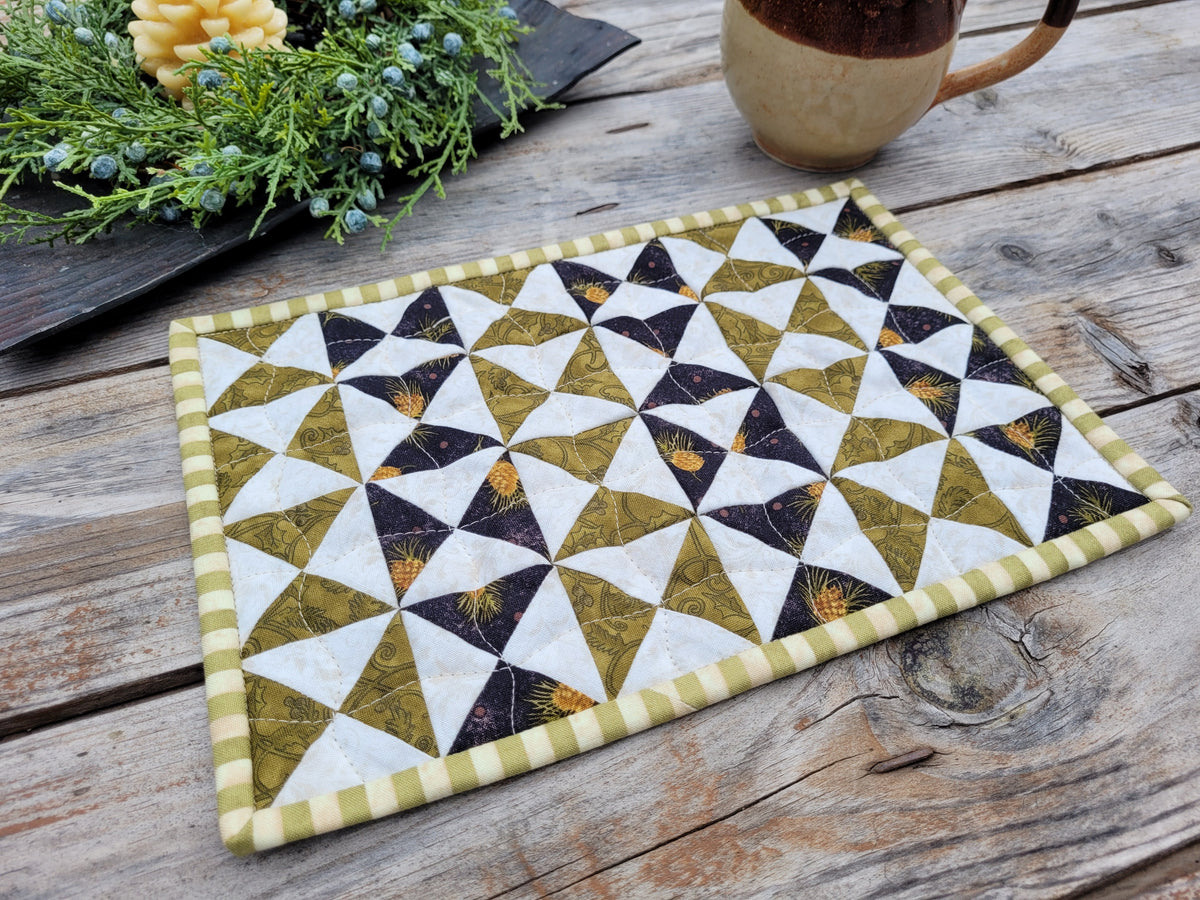 A small rectangular quilt with a patchwork triangles in stripes of olive green and black on an off-white background, featuring a green and cream striped border, displayed on a rustic wooden table next to a ceramic mug and greenery.