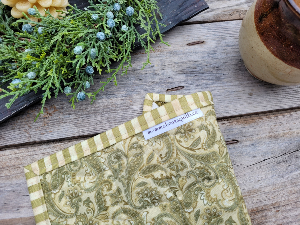 A small quilt is folded to show the backing of a green and beige paisley pattern and striped border, laid on a rustic wooden surface with decorative greenery and a ceramic mug nearby. A small label reads 'momma bears quilts'.