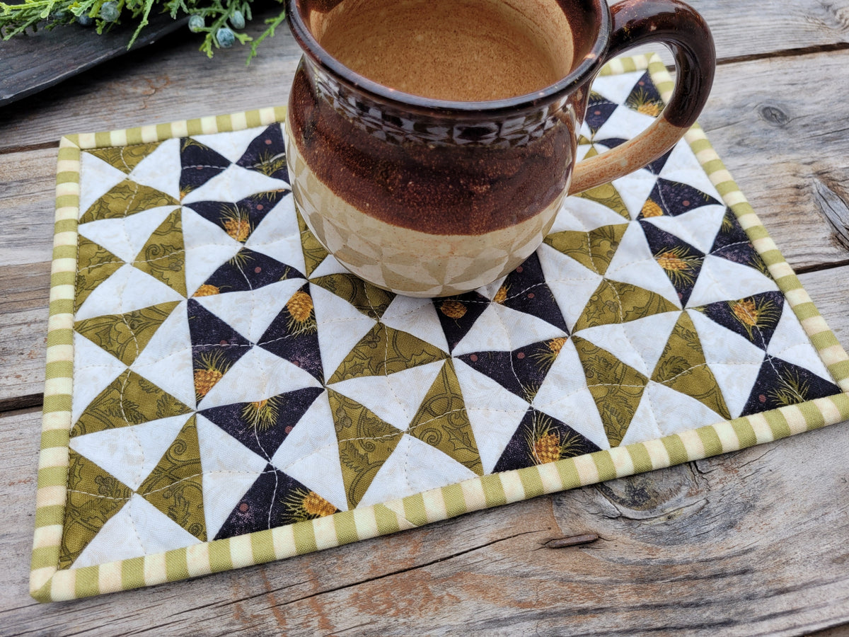 A small quilted fabric placemat with a geometric triangle pattern in olive green, white, and black, featuring a striped border, shown on a rustic wooden table with a ceramic mug placed on top.