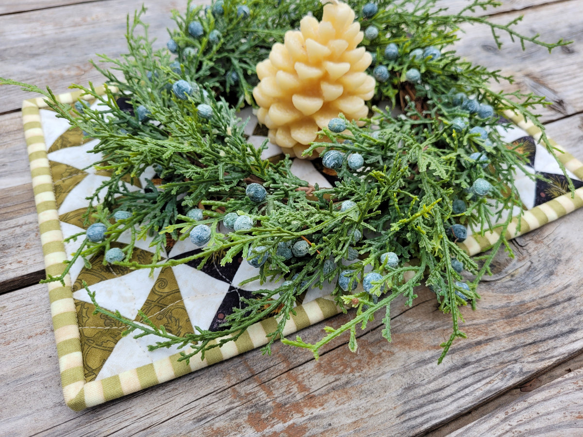 A rectangular quilted placemat with a geometric triangle pattern in olive green, cream, and black, styled with green cedar branches, blue berries, and a yellow candle on a rustic wooden table.