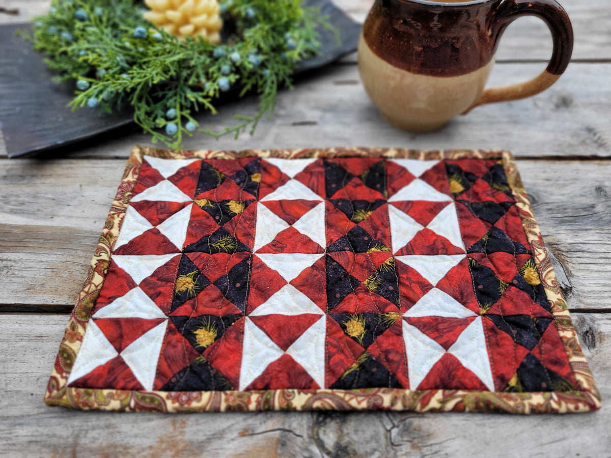 A red and black mini quilt is shown on a weathered wooden table with greenery, a candle and mug partially visable across the top of the photo.