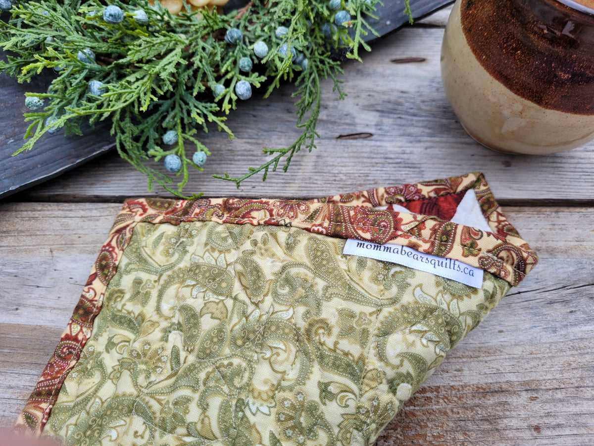 A small quilt is folded to show the backing, on a weathered wooden table with greenery and a mug partially visable.