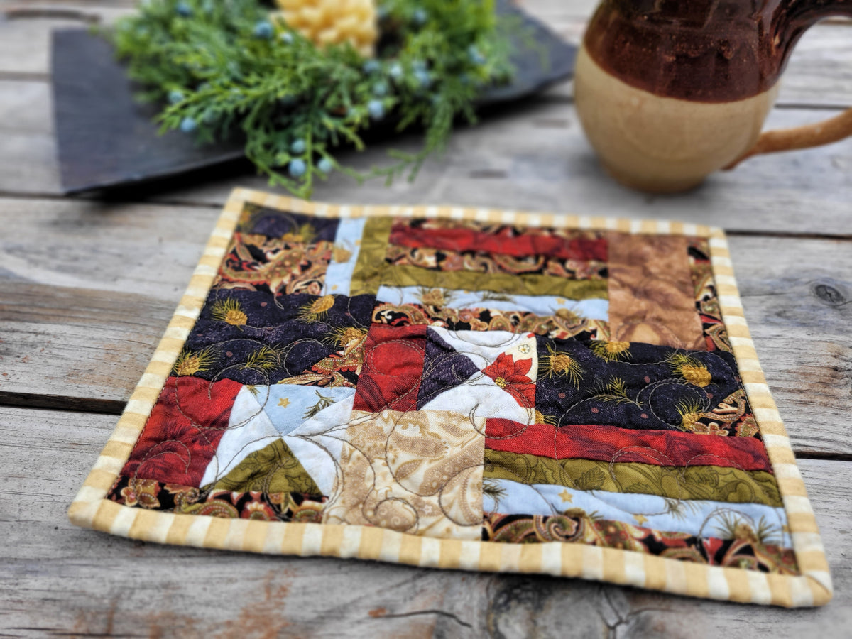 A square quilted mug rug with a patchwork design featuring rustic holiday fabric, finished with a yellow-and-white striped border, displayed on a weathered wooden table.