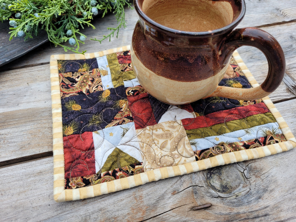 A square quilted fabric coaster with a rustic patchwork design and striped border, shown on a weathered wooden table with a ceramic mug resting on top.