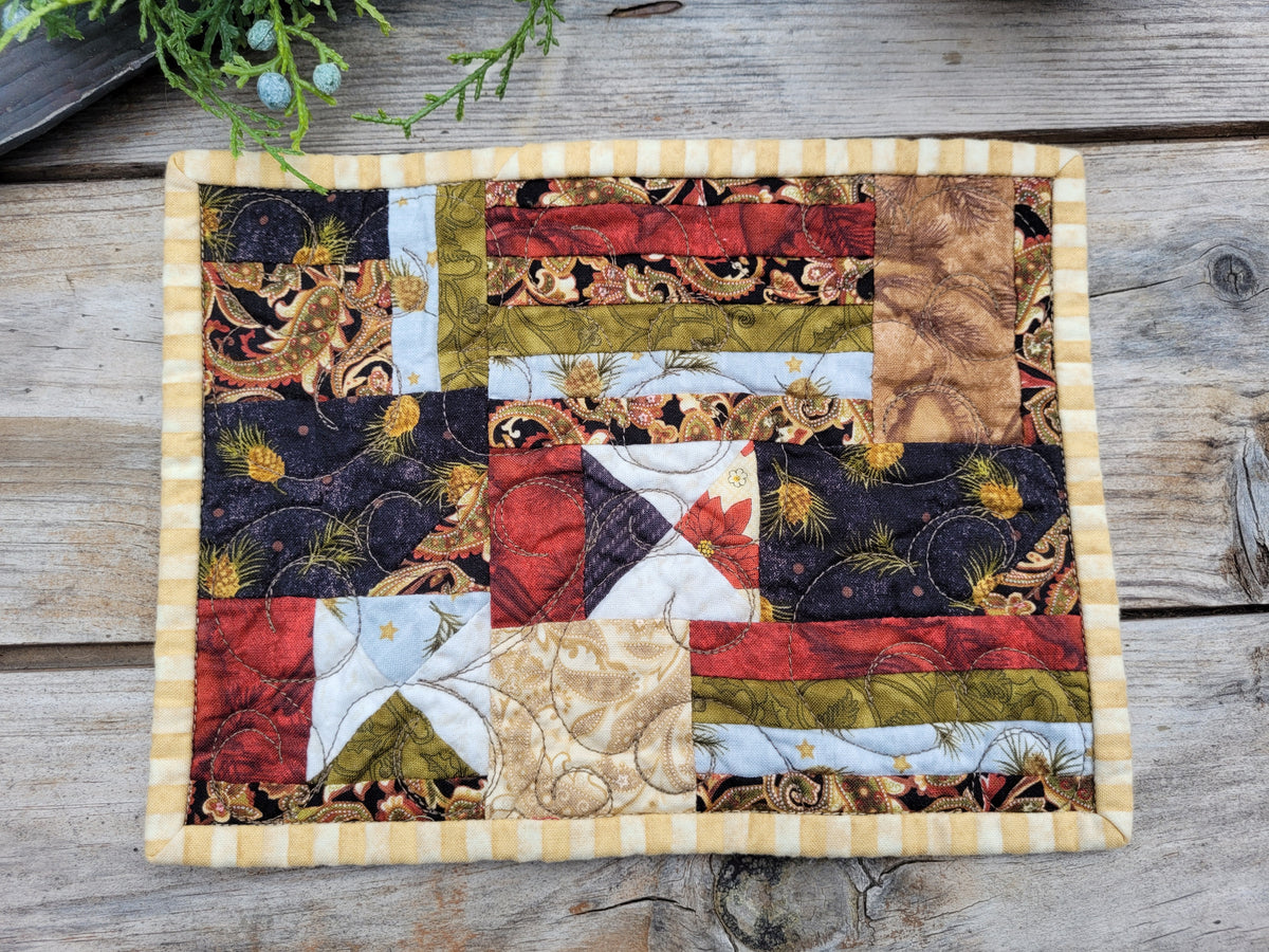 A square quilted fabric mug rug with a patchwork design featuring rustic red, green, beige, and black patterns, finished with a yellow-and-white striped border, displayed on a weathered wooden surface.
