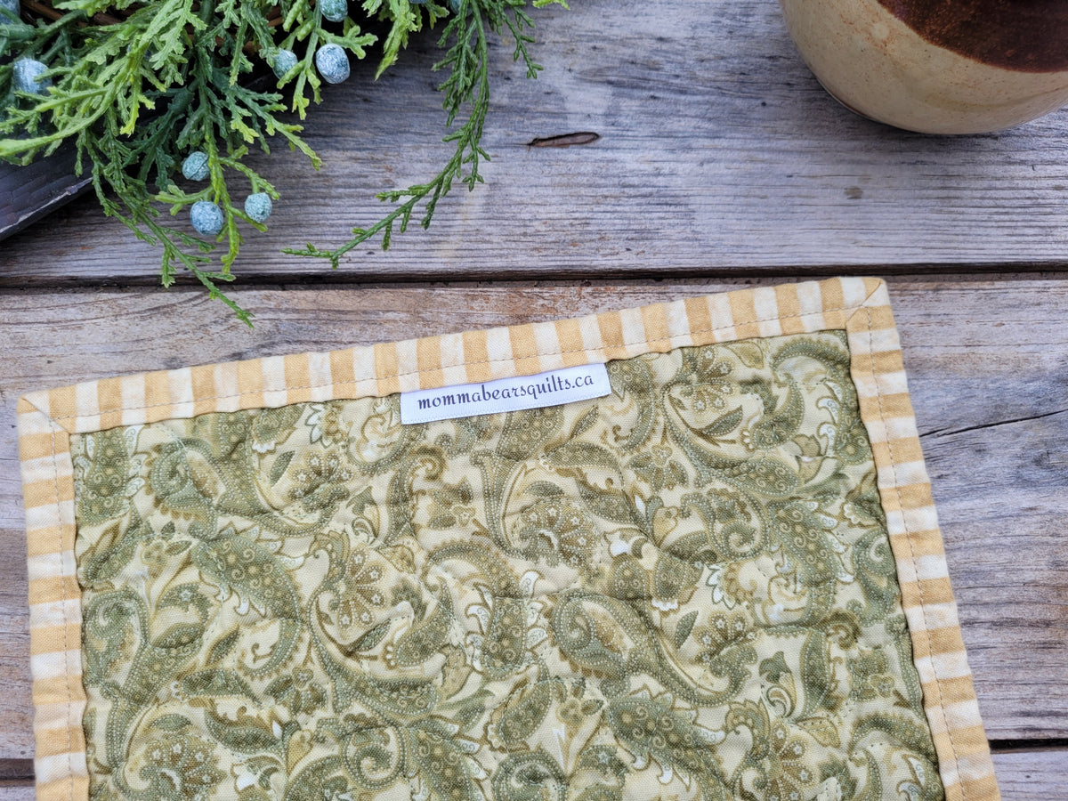 Showing the back of the mug mat with a green and beige paisley pattern and a yellow-and-beige striped border, laid on a rustic wooden surface. 