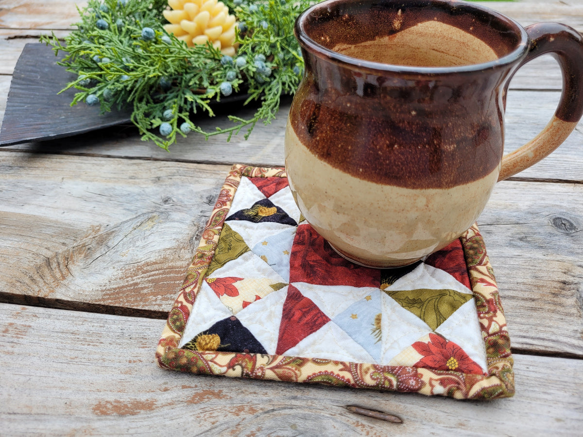 A handmade ceramic mug with a brown and beige glaze sits on a square quilted fabric coaster with a floral and geometric patchwork pattern, placed on a rustic wooden table.