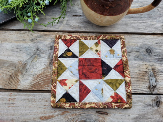 A square quilted fabric trivet or coaster with a patchwork starburst design, placed on a rustic wooden tabletop next to a ceramic mug and greenery.