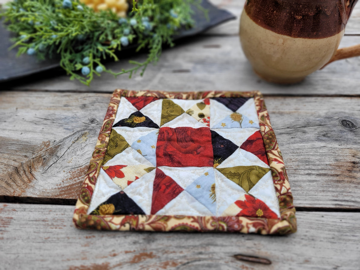 A square quilted fabric coaster with a geometric starburst pattern in red, cream, black, olive green, and floral prints, set on a rustic wooden table next to a ceramic mug.