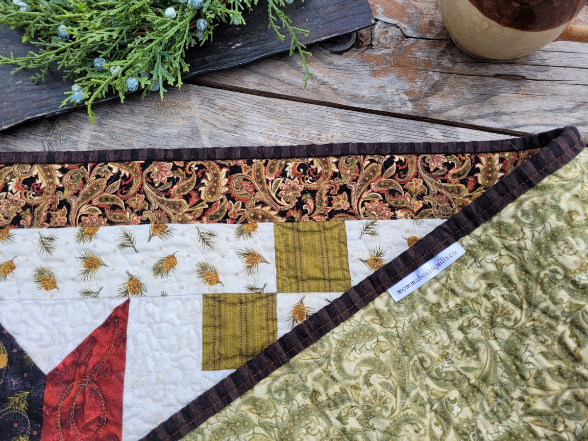 Christmas table runner is folded to show backing with a brand lable along the binding edge, styled on a weathered wooden table with a mug and greenery partially visable along the top.