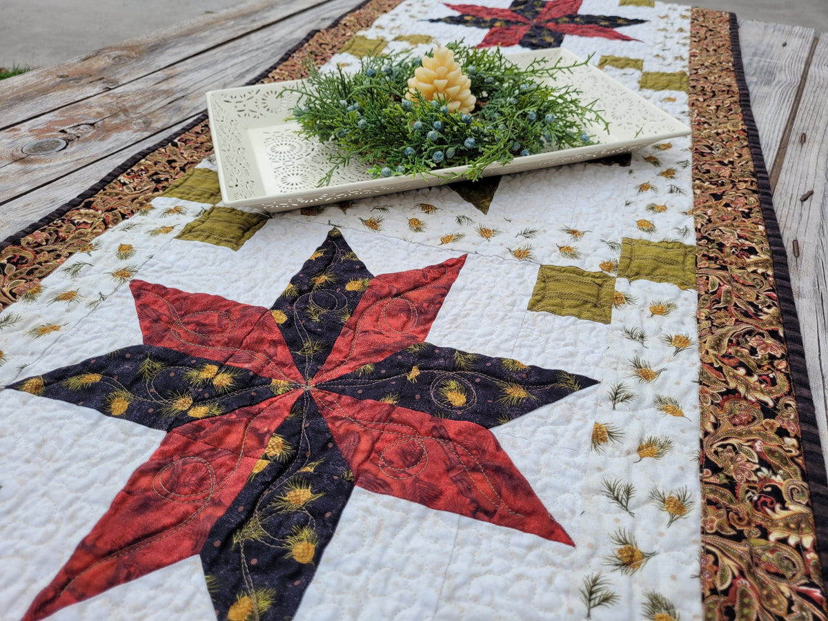 A handmade quilted table runner laid out on a rustic wooden table, featuring a large red and black star motif, a white floral border, and a decorative white tray with greenery and a candle on top.