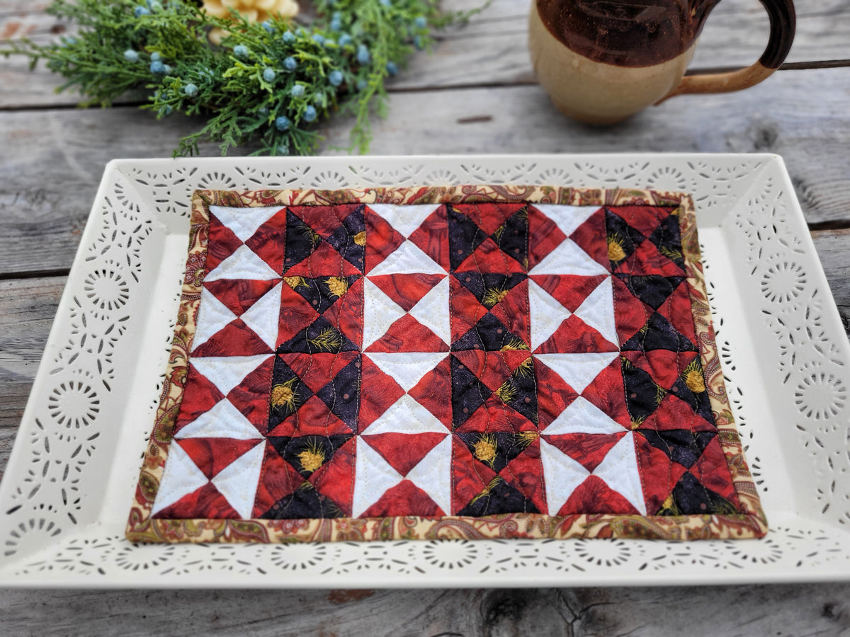 A striking mini quilt with black and red patchwork stripes is shown on a white decorative tray with greenery and a mug in the background. 