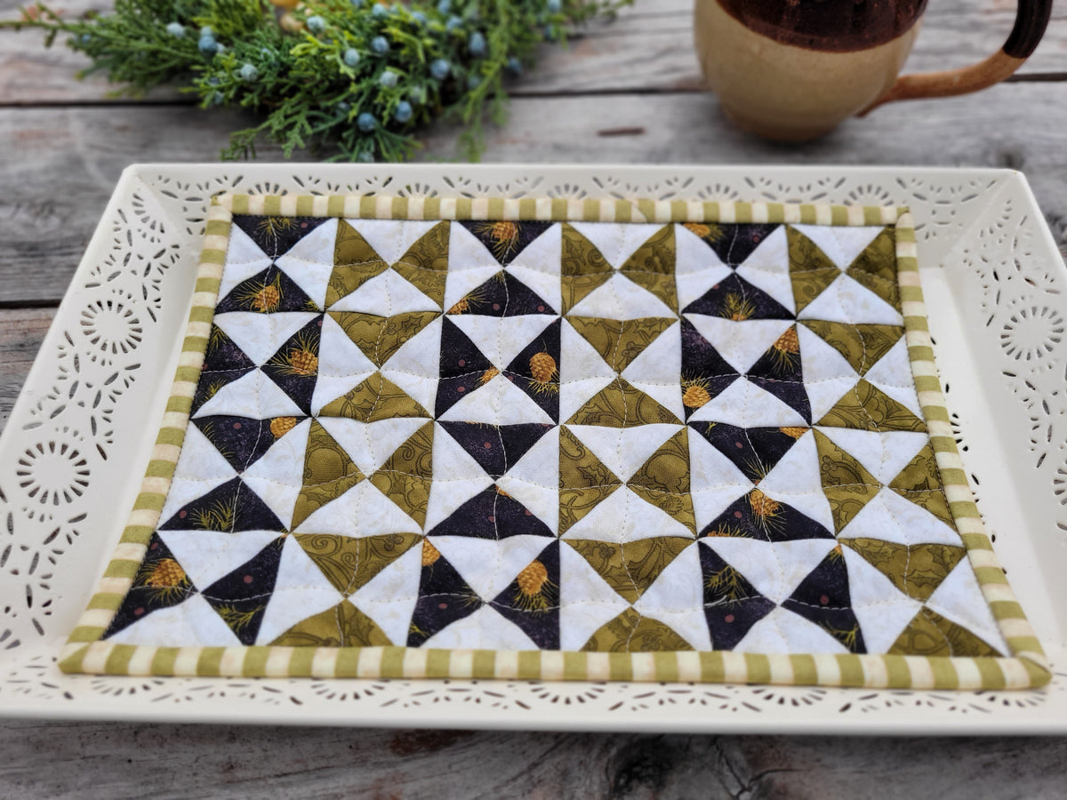 A square quilted fabric placemat with a geometric triangle pattern in olive green, white, and black, displayed on a white decorative tray on a rustic wooden table.