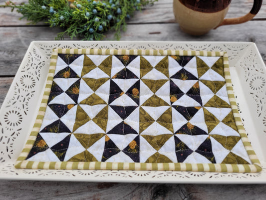 A square quilted fabric placemat with a geometric triangle pattern in olive green, white, and black, displayed on a white decorative tray on a rustic wooden table.