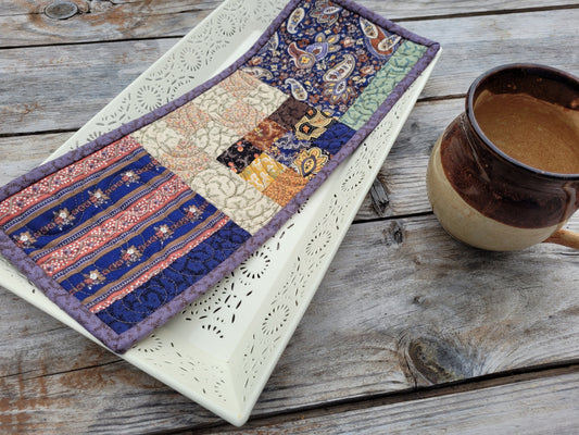 A rectangular patchwork mini quilt with a purple border and multicolor fabric panels, displayed on a white ceramic tray next to a brown ceramic mug on a rustic wooden table.