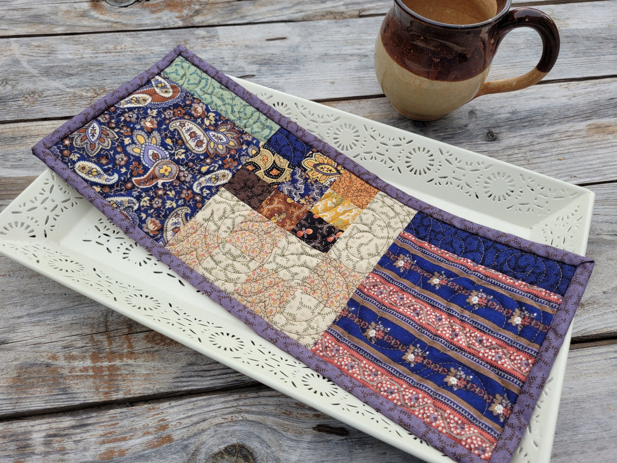 A small rectangular patchwork quilt with a multicolor patterned design, resting on a white ceramic tray with a rustic wooden table and a ceramic mug in the background.