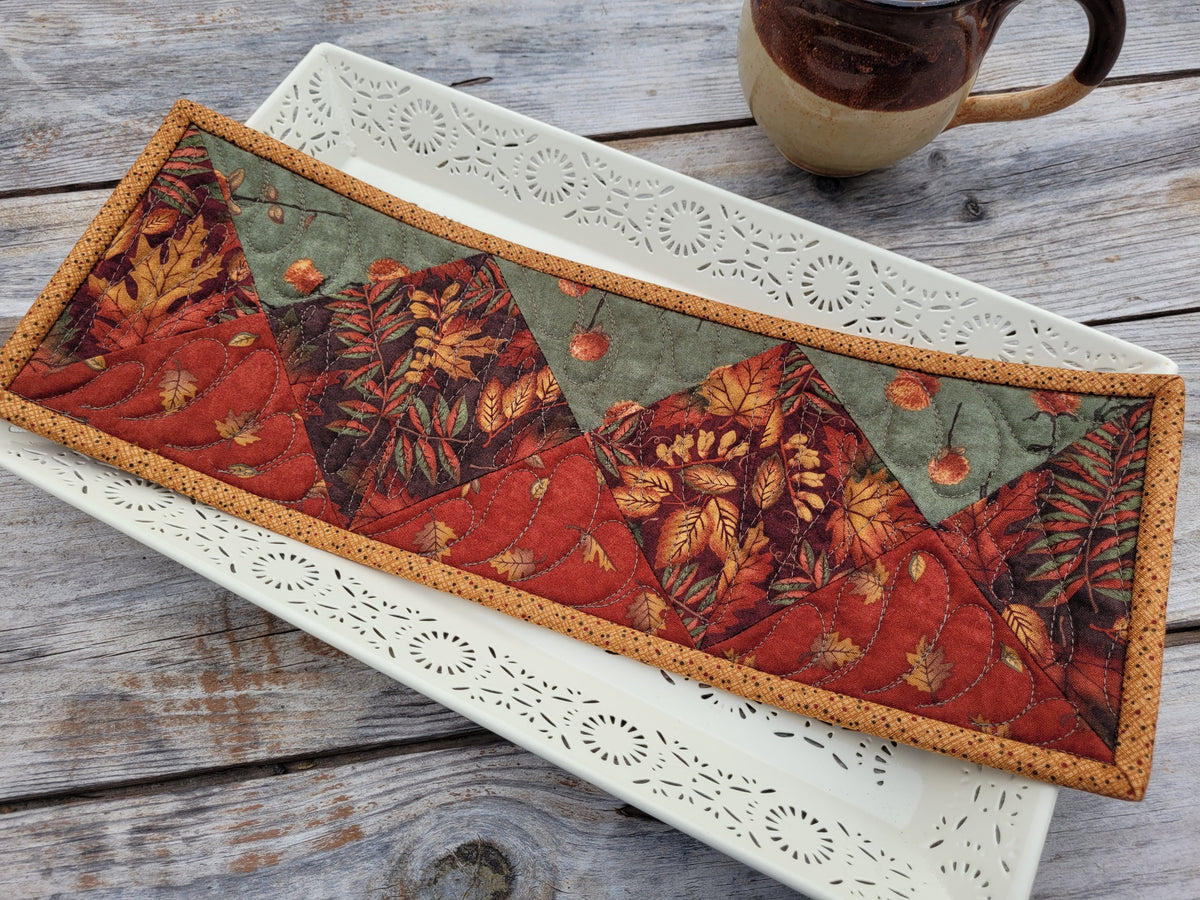 An autumn-themed skinny mini quilt with geometric patterns in red, orange, brown, sage green and a tan border. The quilt is displayed on a white decorative tray over a rustic wooden table, with a ceramic mug partially visible in the top right corner.