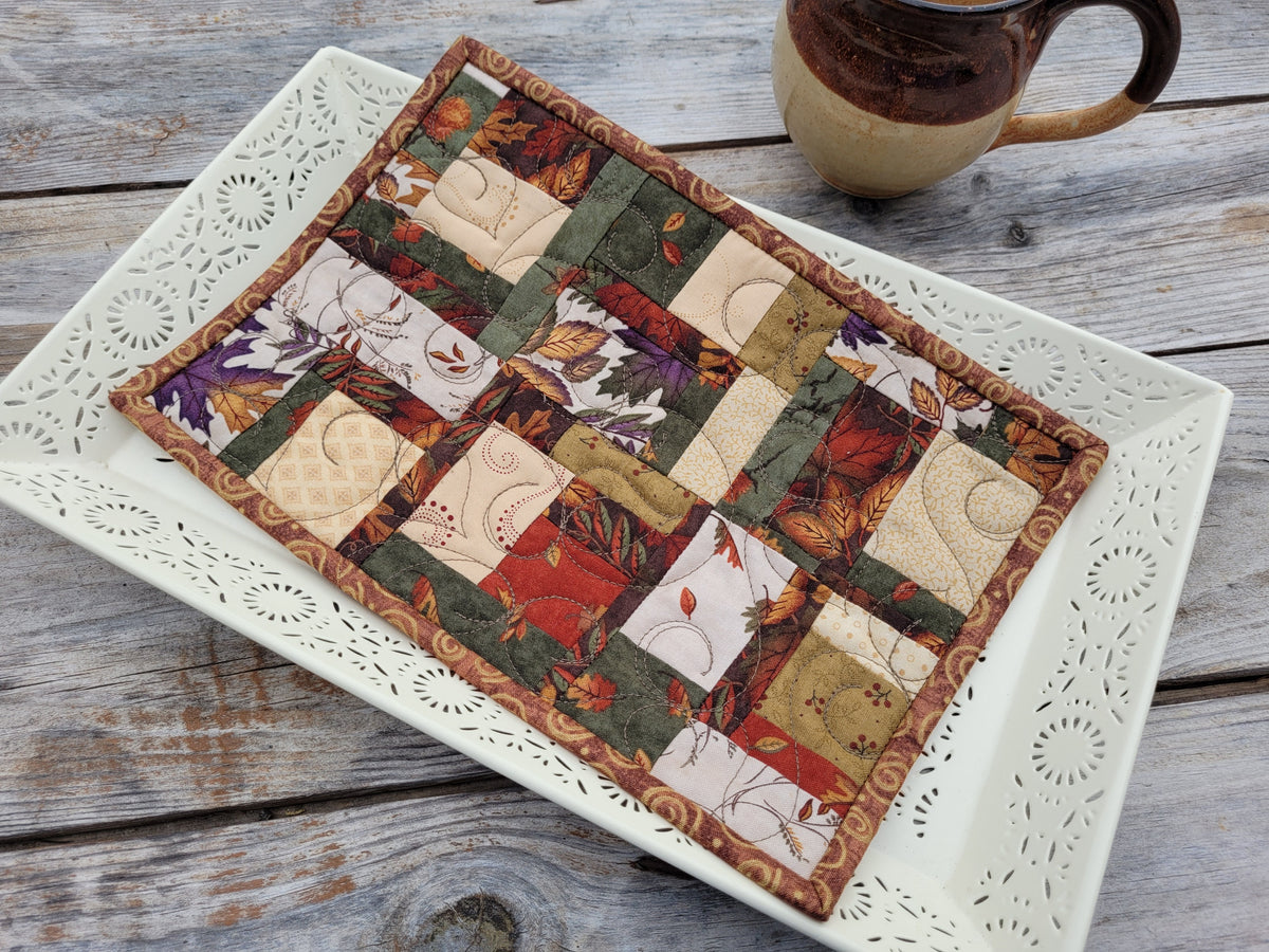 A small rectangular patchwork quilt with an autumn leaf and floral pattern, displayed on a white decorative tray on a rustic wooden table next to a ceramic mug.