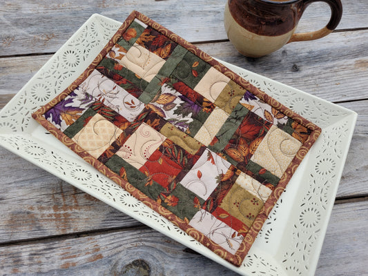 A small rectangular patchwork quilt with an autumn leaf and floral pattern, displayed on a white decorative tray on a rustic wooden table next to a ceramic mug.