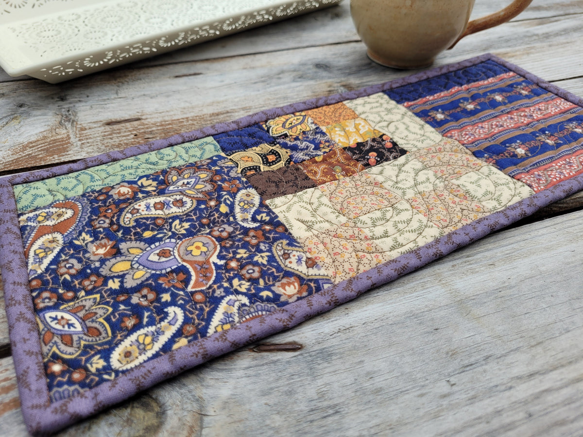 A small rectangular patchwork quilt with a purple border, featuring various fabric panels with paisley, floral, and striped patterns in purple, beige, orange, and brown, styled on a rustic wooden tabletop next to a ceramic mug.