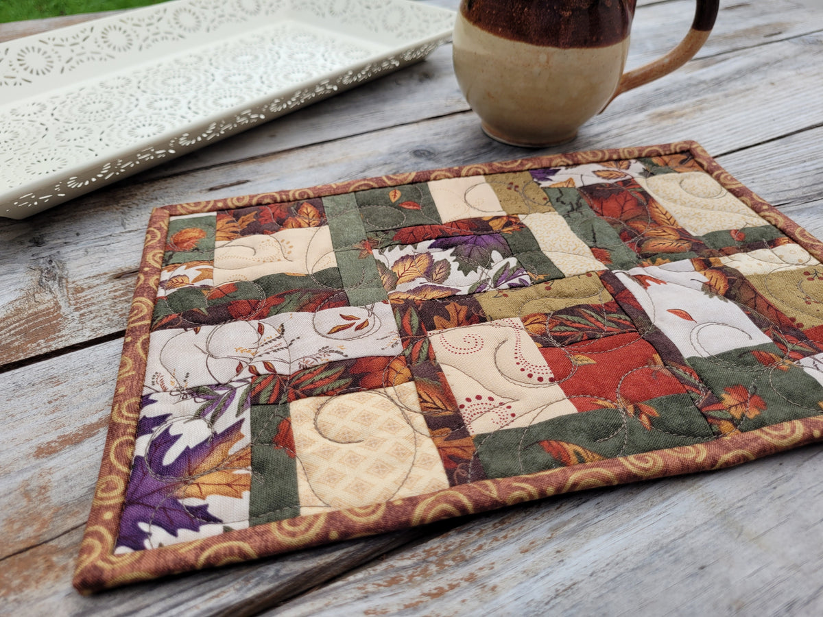 A square quilted fabric placemat with a patchwork design featuring autumn leaves and floral motifs in earthy tones, styled on a rustic wooden table next to a ceramic mug and a white decorative tray.