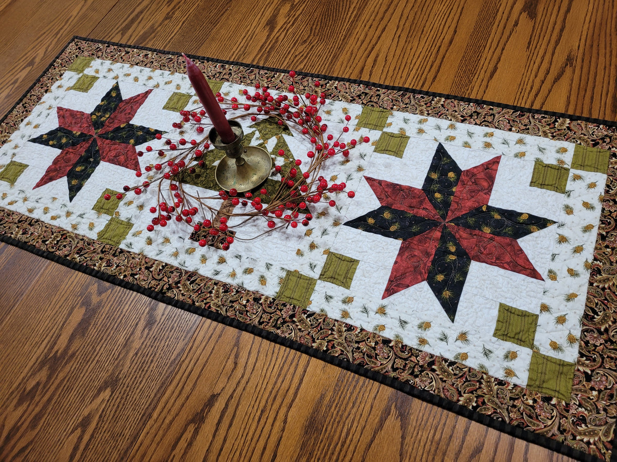 A rectangular quilted table runner with a rustic brown patterned border, featuring two large red and black star motifs on a white background with a green patchwork tree partially visable in the centre. The runner is styled on a wooden table with a red candle and red berries in a brass holder, which is sitting on the patchwork tree in the centre of the runner.