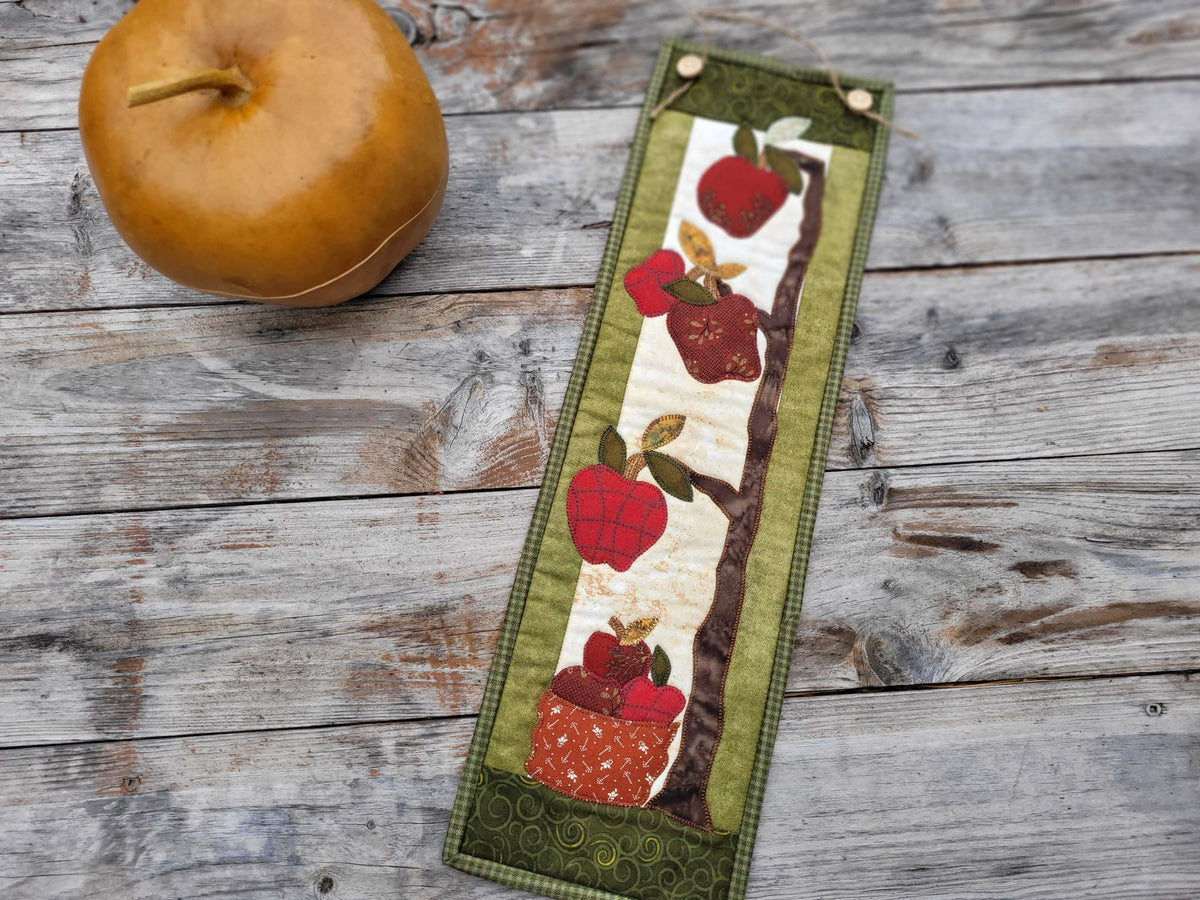 A skinny apple themed wall quilt is laid on a weathered wood table with a golden apple-shaped gourd nearby.