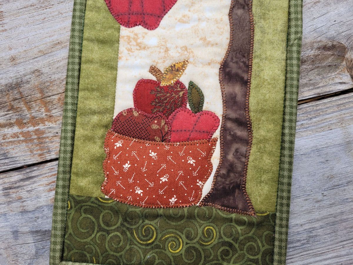A close up of the bottom portion of the narrow wall quilt is shown, featuring applique shapes of a basket filled with red apples and a partially visable tree.