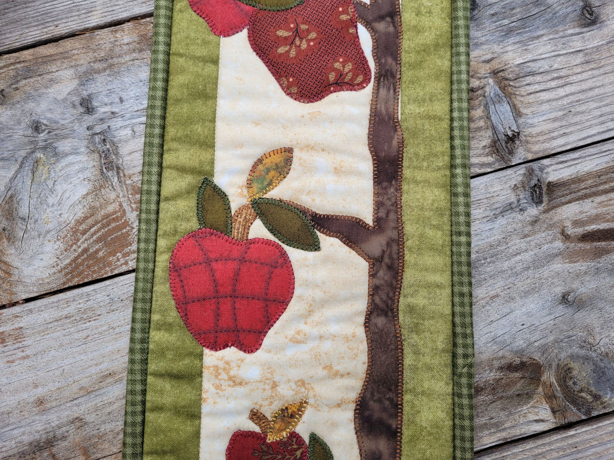 Close up of a narrow quilt showing red applique apples with leaves and a tree and green borders,