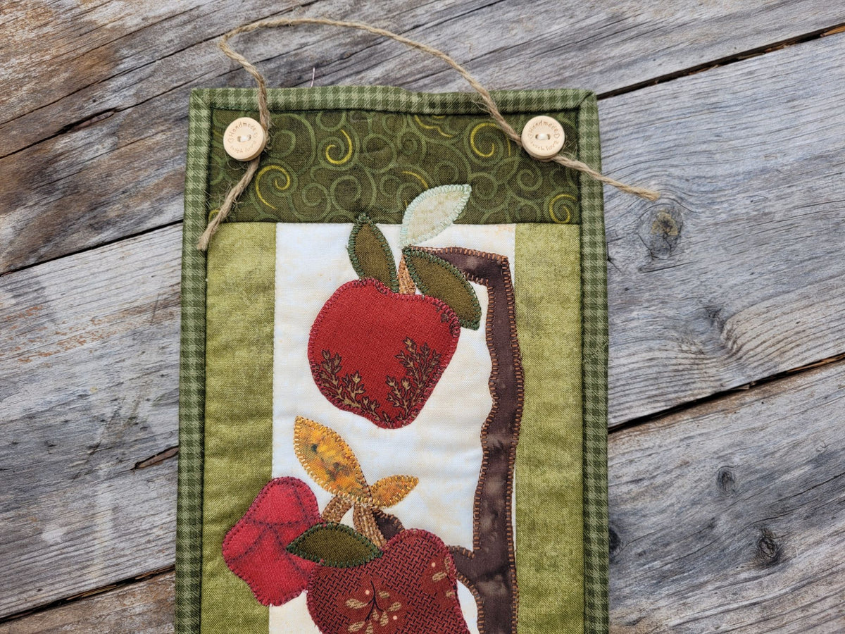 Close up of a narrow apple themed wall quilt showing the two wooden buttons holding the jute hanging string and red applique apples, leaves and tree branches.