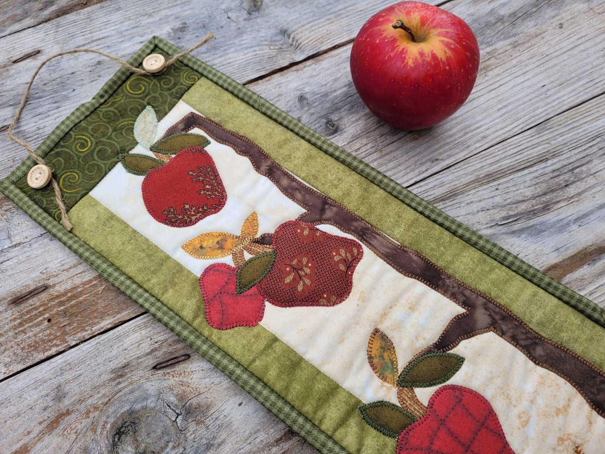 Close up of a narrow apple themed wall quilt is partially visable showing red applique apples, leaves, tree branches, and green borders. Shown on a weathered wood surface with a red apple nearby.