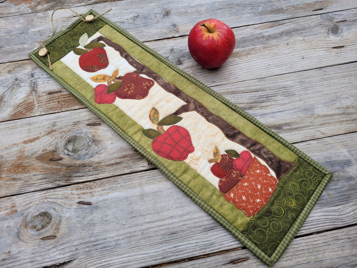 A skinny apple themed wall quilt is laying diagonally on a weathered wooden table with a red apple nearby.