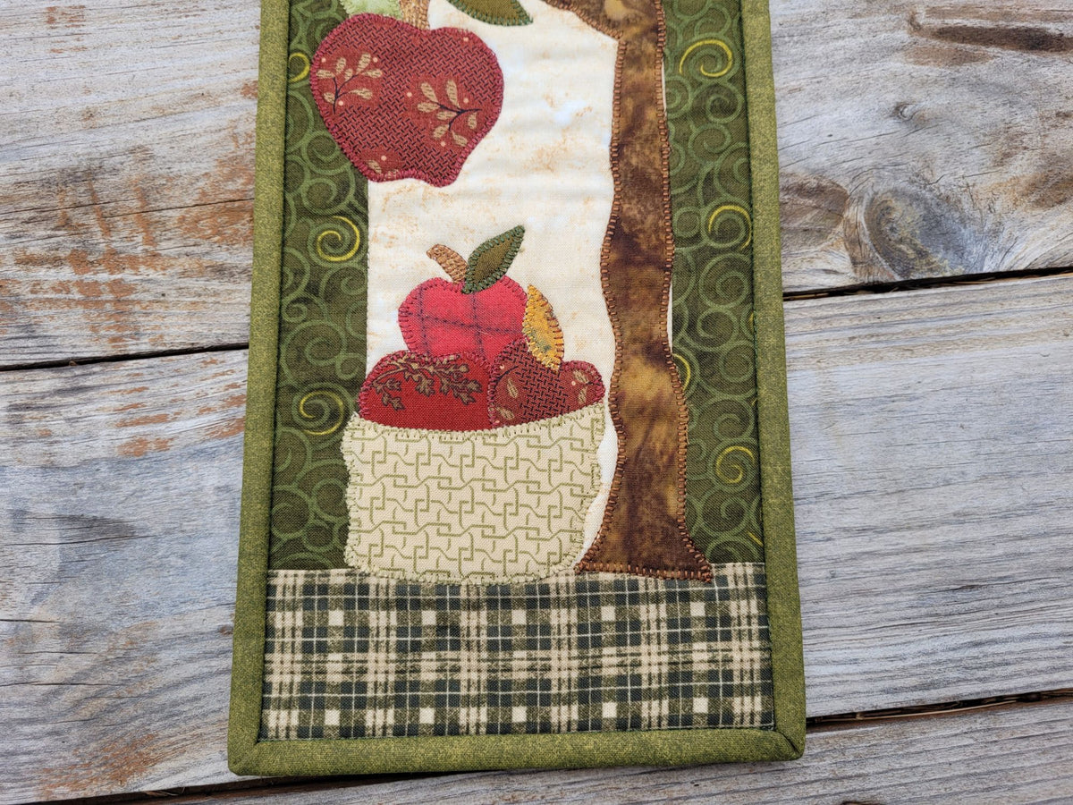 Close up of the bottom portion of the narrow wall quilt, showing the basket of red apples and part of the tree, on a weathered wood surface.