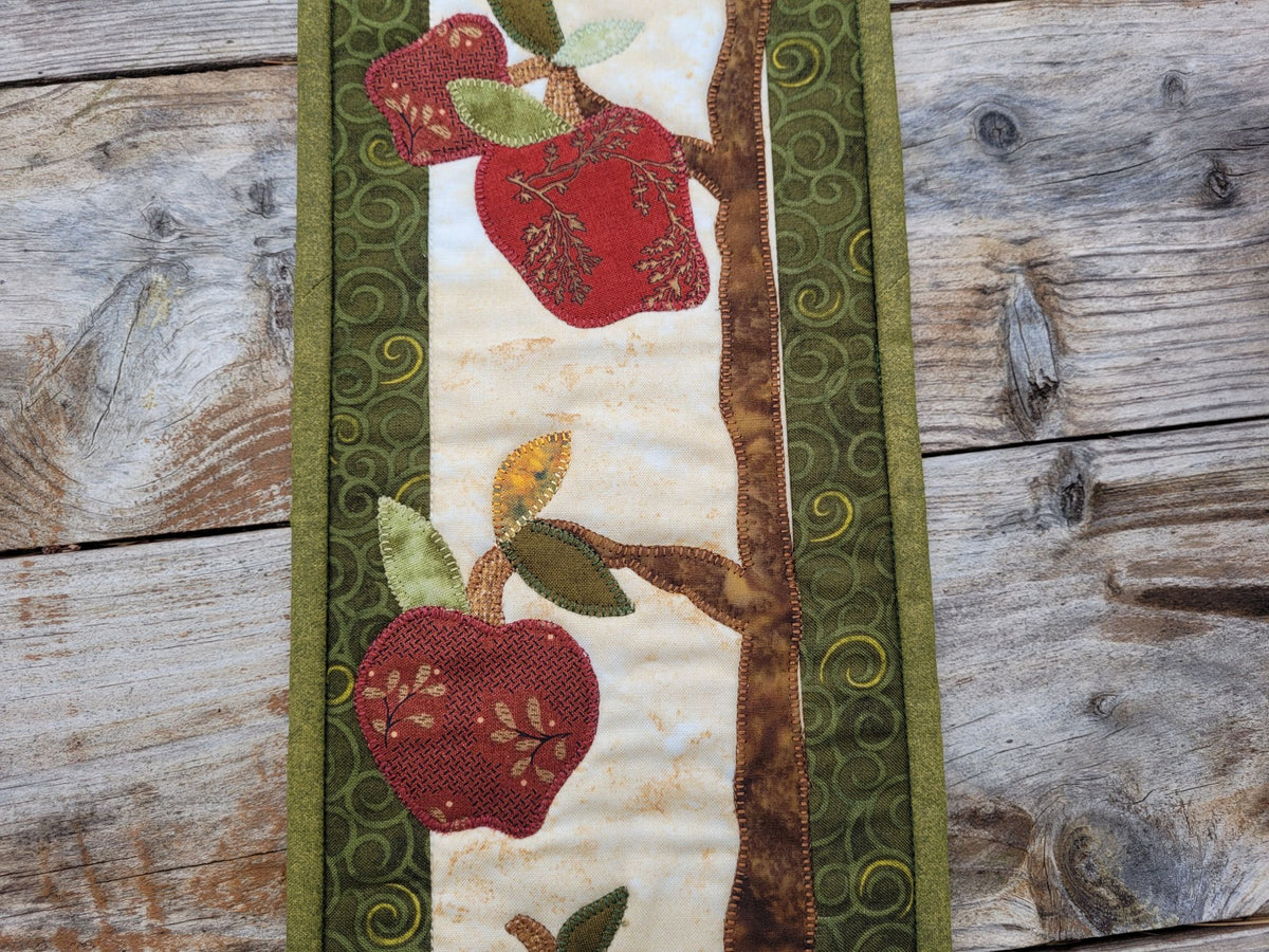 A portion of narrow quilt is shown on a weathered wood surface. The quilt features applique designs of red apples and leaves on tree branches with green patterned borders. 
