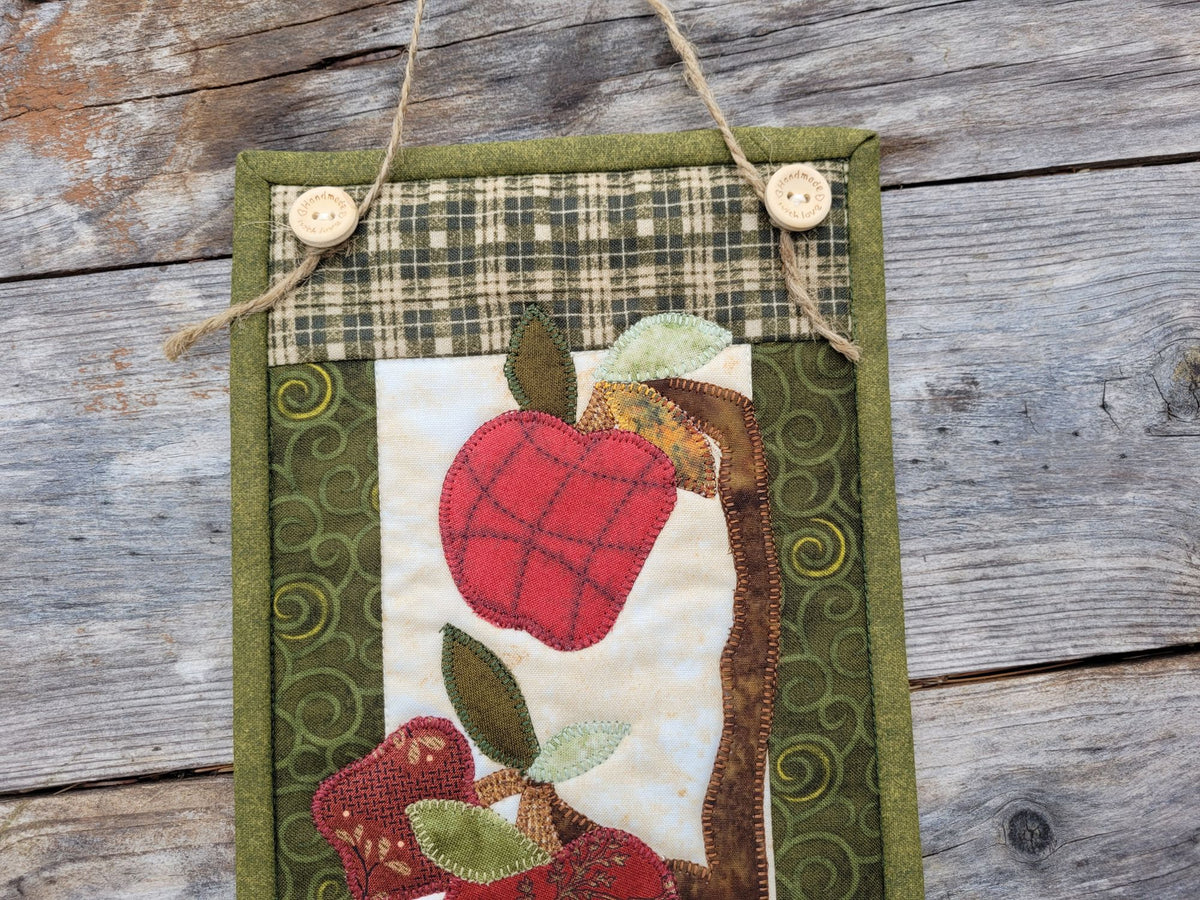 A narrow quilt is partially visable on a weathered wood surface, showing the jute hanging string on two wooden buttons at the top and the applique shapes of red apples and leaves on a tree banch.