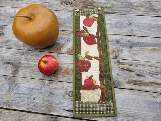 A skinny apple themed wall quilt is shown on a weathered wood table with a red apple and large golden apple-shaped gourd sitting nearby.