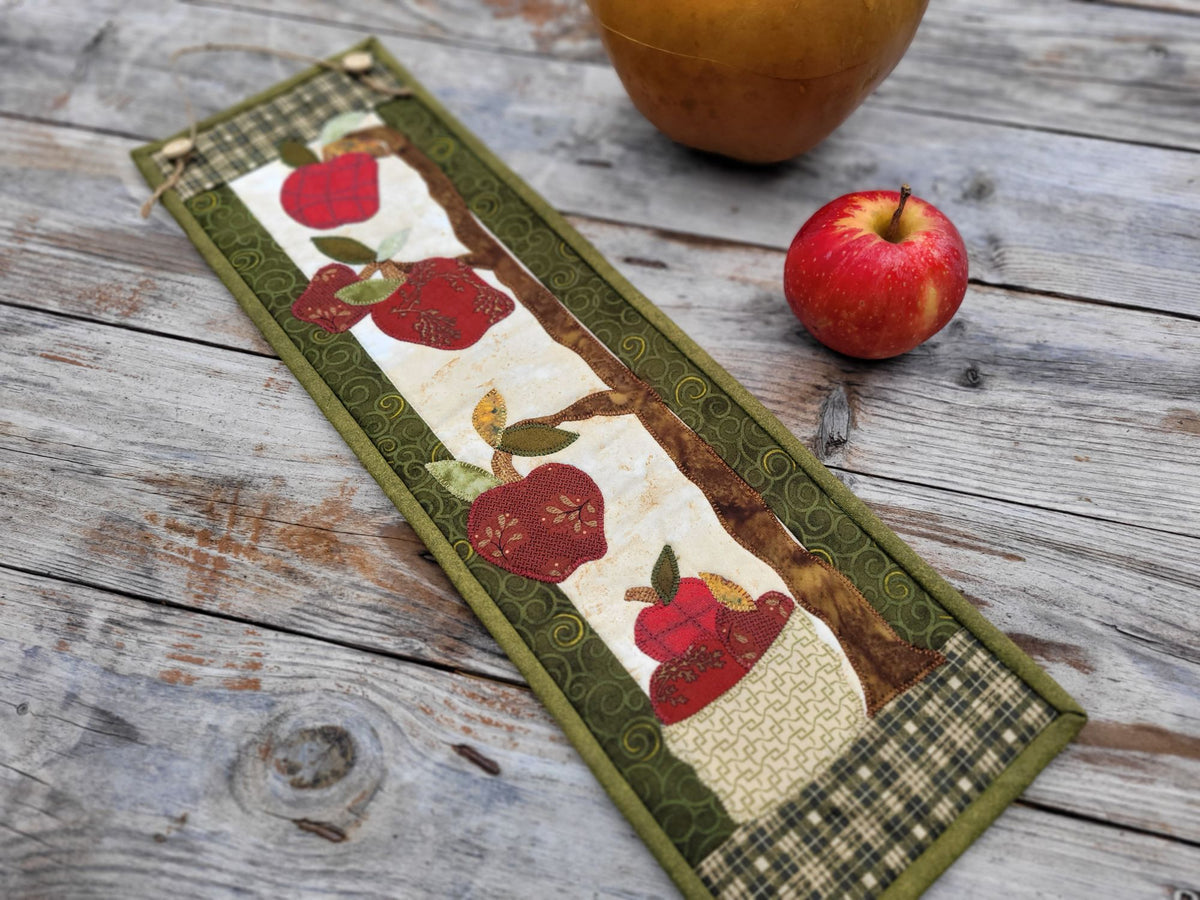 A skinny apple themed wall quilt is shown laying diagonally on a weathered wooden table with a red apple nearby.