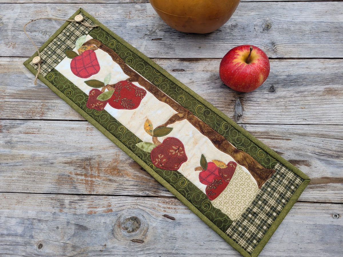 A skinny wall quilt is laying diagonally on a weathered wood table with a red apple nearby and a gourd partially visable. The quilt features applique apples with green patterened borders.
