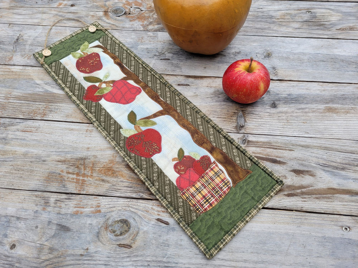 A narrow wall quilt is shown laying diagonally on a weathered wood table with a red apple nearby and a large golden gourd  partially visable. Quilt features red apples with green borders.