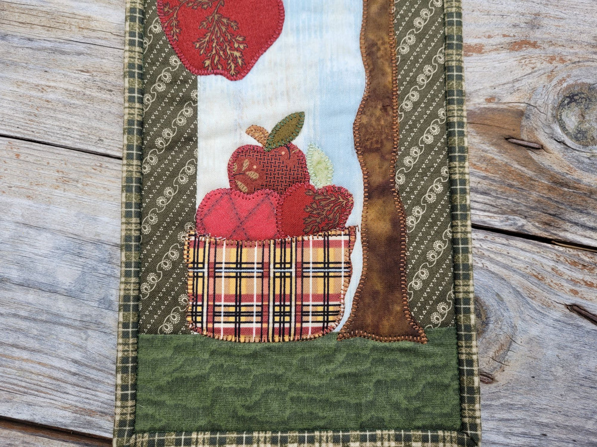 Close up of a narrow quilt partially visable on a weathered wood surface. Quilts shows applique designs of a tree and apples in a basket.