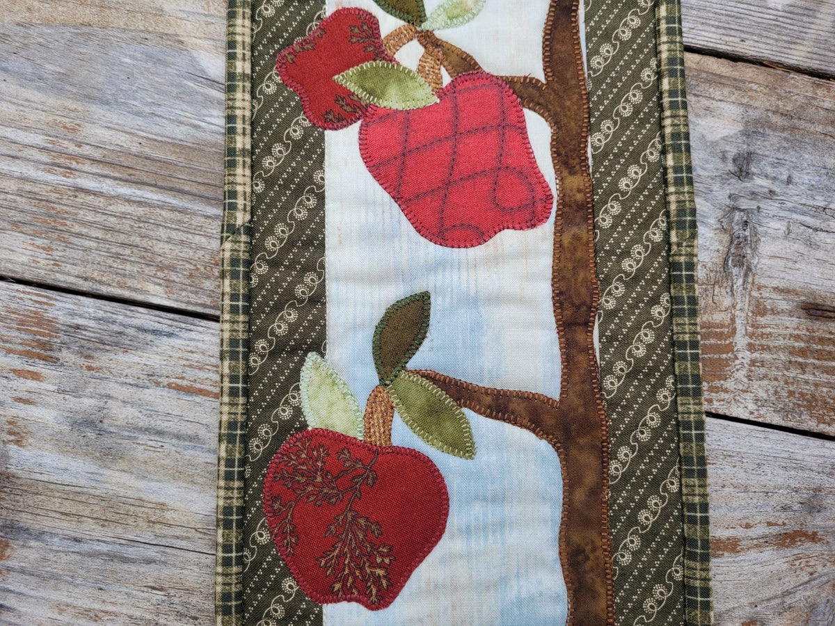 Close up of the middle portion of the skinny wall quilt showing red applique apples and leaves on tree branches and the green patterned borders. Shown on a weathered wood surface.